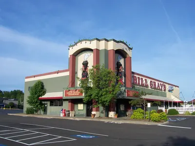 A Bill Gray's restaurant photographed from its own parking lot under a clear blue sky. The building is painted dark green and deep red and has two corner towers, each carrying a raised black steam locomotive with a puff of smoke and a curved BILL GRAY'S sign over the top of it. A long BILL GRAY'S sign in deep red letters runs along the right-hand wall beside a blue script Abbott's Frozen Custard sign, and red panels under the awnings read HOME OF THE WORLD'S GREATEST CHEESEBURGER. Young trees and shrubs stand along the front of the building and the marked spaces of an empty parking lot run across the foreground.
