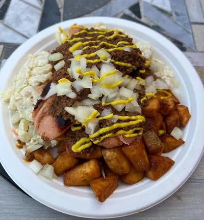 A plate on a mosaic table top: two split red hot dogs lying on chunky home fries and macaroni salad, covered with crumbled brown meat sauce, a scatter of diced raw onion and a zigzag of yellow mustard.