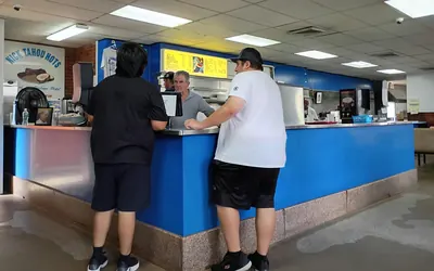 Inside Nick Tahou Hots in 2025. Two customers stand with their backs to the camera at a long blue counter under strip lighting; a member of staff faces them across it. A yellow menu board hangs overhead and a sign on the left wall reads NICK TAHOU HOTS above a drawing of a plate.