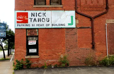The corner of Nick Tahou Hots' red brick building. A hand-painted board high on the wall carries a Coca-Cola logo and reads NICK TAHOU, PARKING IN REAR OF BUILDING, with a green arrow. Below it a boarded-up window carries a LOTTO sign. A Rochester Heritage Trail marker stands at the curb on the left.