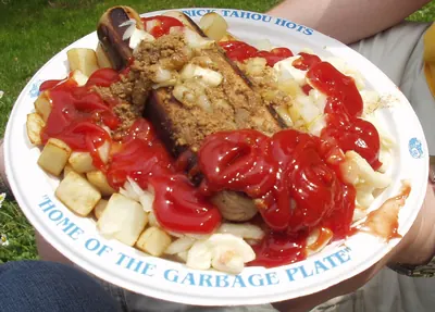 A Garbage Plate held on somebody's lap outdoors on grass. The plate's blue rim reads NICK TAHOU HOTS along the top and, along the bottom, HOME OF THE GARBAGE PLATE. On it are a split red hot dog, home fries, macaroni salad, meat sauce, chopped onion and a heavy pour of ketchup.