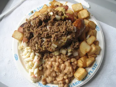 A Garbage Plate seen from above on a plate whose blue rim reads NICK TAHOU and, along the bottom, HOME OF THE GARBAGE PLATE. Baked beans, macaroni salad and chunky home fries are arranged around the edge; a mound of crumbled meat sauce and chopped onion sits in the middle over a split red hot dog.