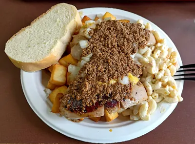 A Garbage Plate at Nick Tahou Hots: two split white hot dogs lying across home fries and macaroni salad, buried under a layer of crumbled brown meat sauce with chopped raw onion and a line of yellow mustard through it. Two slices of white Italian bread lie on the rim of the plate and a fork rests on the right.