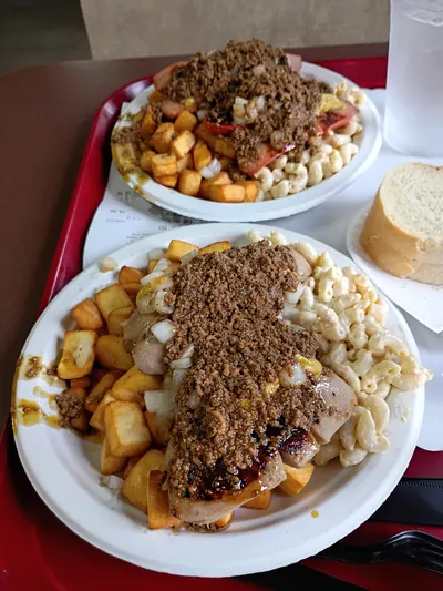 Two Garbage Plates on a red cafeteria tray, one behind the other. The plate at the back carries split red hot dogs under the meat sauce; the one at the front carries pale white hots. Both sit on the same home fries and macaroni salad. A slice of Italian bread on its own plate and a cup of water stand beside them.