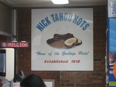 A large painted sign on the brick wall inside Nick Tahou Hots. It reads NICK TAHOU HOTS in blue over a drawing of a steaming plate of hot dogs, beans and bread, then in script "Home of the Garbage Plate", then Established 1918 in red. A red LED display on the left reads 5 MILLION.