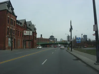 Looking east along West Main Street in Rochester, NY on an overcast day. On the left stands a long red brick building with steep gables and tall arched windows, a hanging sign on it reading NICK TAHOU. A green railroad bridge crosses the road ahead and downtown towers stand behind it.