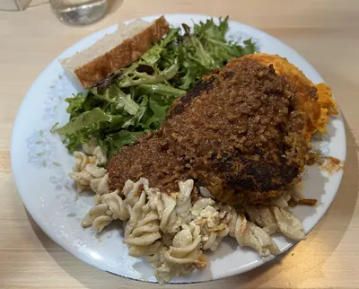 A Compost Plate on a floral-rimmed china plate: a browned patty under a thick shredded sauce, sitting on a bed of macaroni salad, with a heap of green salad leaves, a slice of bread and a scoop of orange sweet potato around it.